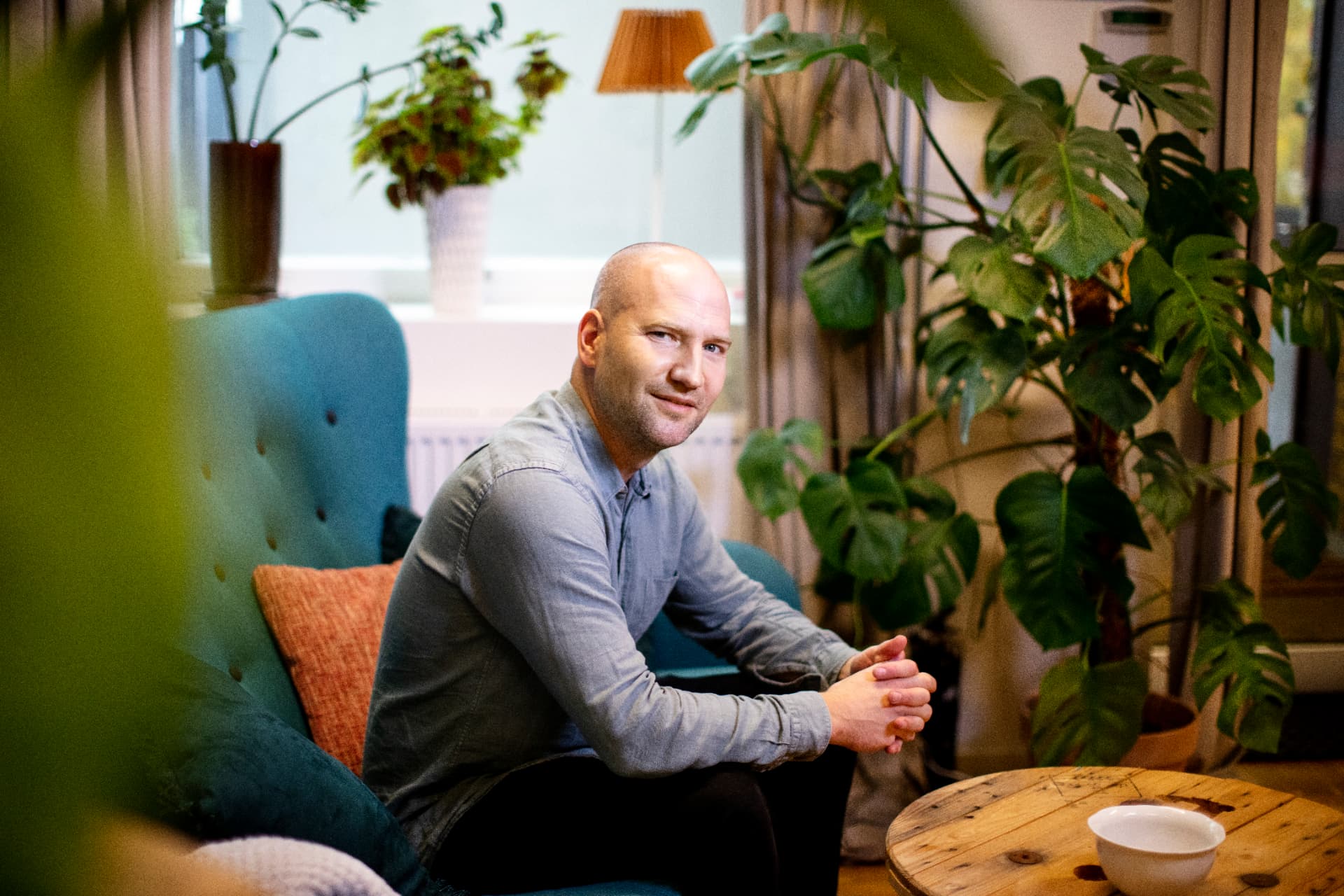 Simon Strandvik sitter i en blågrön fåtölj bland gröna växter i ett soligt vardagsrum och ler mot kameran